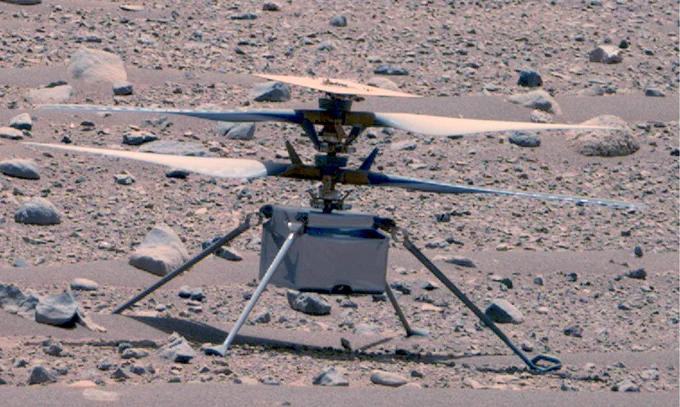 A color photo of the Ingenuity Mars helicopter shows the rotorcraft sitting on the surface of the Red Planet. Its four slanted legs rest on the rocky surface supporting the box-shaped main body and double propellers. The helicopter takes up the majority of the image, and the rest of the frame is speckled with reddish brown rocks and pebbles. The image was taken by the Perseverance rover’s Mastcam-Z on April 16, not long after the rotorcraft’s 50th flight.