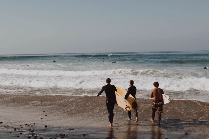 Image of 3 surfers walking on the beach towards the water. The waves are rolling in and we see other surfers in the background.