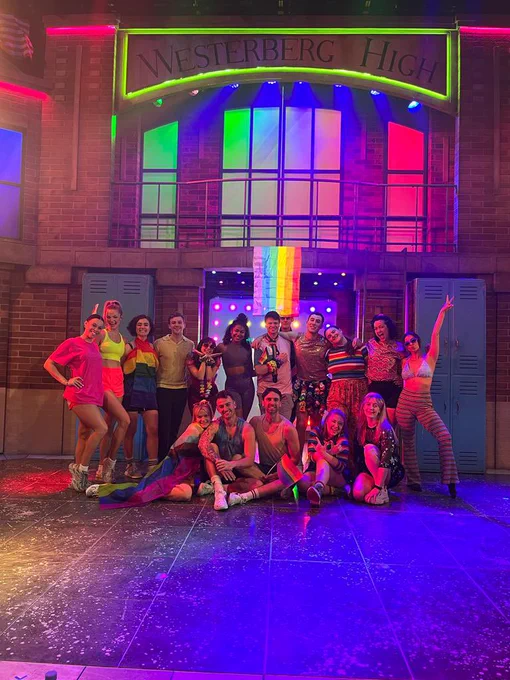 The cast of Heathers The Musical in London, onstage with lights set to rainbows - all smiling and looking into camera.