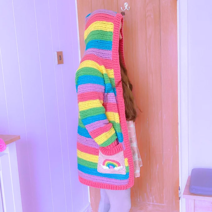 A woman with long brown hair is shown wearing a pastel rainbow striped hooded cardigan with baby pink pockets that feature pastel rainbows with white clouds at both ends
