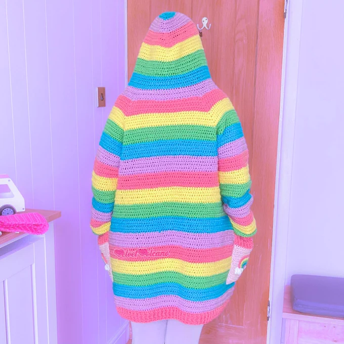 A woman with long brown hair is shown wearing a pastel rainbow striped hooded cardigan with baby pink pockets that feature pastel rainbows with white clouds at both ends