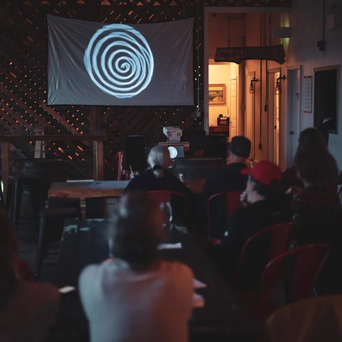Color photograph of a crowd of people watching a film that is projected on a screen hanging on a wall.
