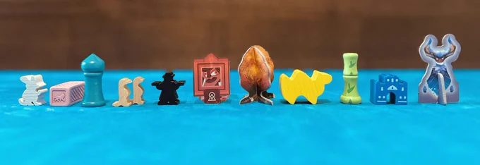 Board game components arranged in the Pride Flag order. A white field mouse, pink brick, cyan tower, brown meerkat, black gunslinger, red picture, orange tree, yellow camel, green bamboo, blue palace, and violet creature