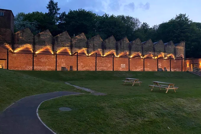 Warm lights shining on the red brick wall of the Mills' colonnade at dusk. In front is a patch of grass with 2 wooden picnic benches in its centre.