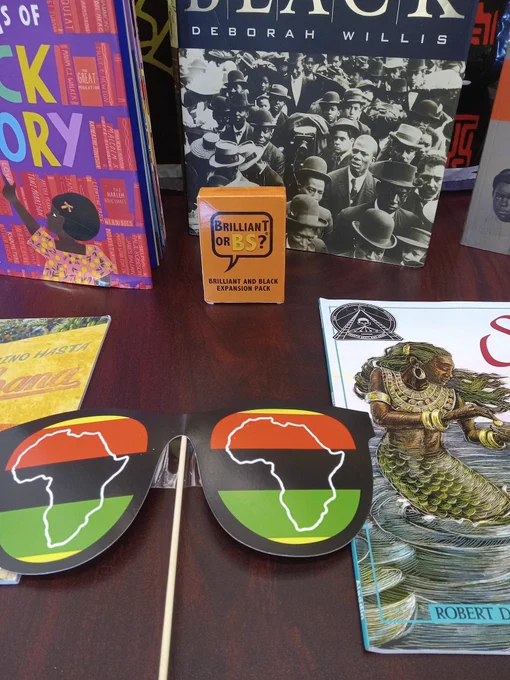 Photo of Afrocentric display on a table. Books featured include The ABCs of Black History by Rio Cortez, illustrated by Lauren Semmer;  Reflections in Black by Deborah Willis; Todo el Camino Hasta La Habana by Margarita Engle, illustrated by Mike Curato; Sukey and the Mermaid by Robert D. San Souci, illustrated by Brian Pinkney; and Pictures with Purpose and a souvenir book by the National Museum of African American History and Culture. The display also contains a Brilliant or BS card game and oversized sunglasses in the red, black, and green colors of the Pan-African flag. 