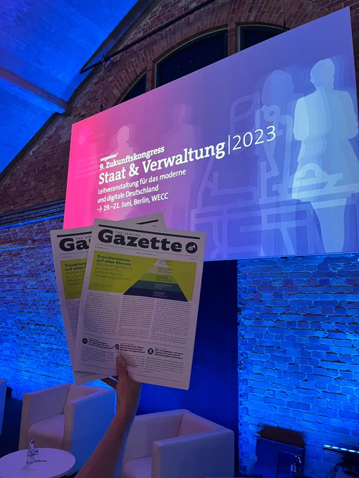 Jemand hält zwei Zeitungen hoch vor einem großen Bildschirm, auf dem steht neunter Zukunftskongress, Staat und Verwaltung 2023