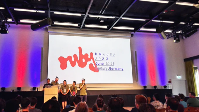 A photo of a stage ath the ruby unconf. 5 People (the organziers) are standing in front of a huge presentation canvas where it's written: Ruby Unconf 2023, June 10-11, Hamburg, Germany
