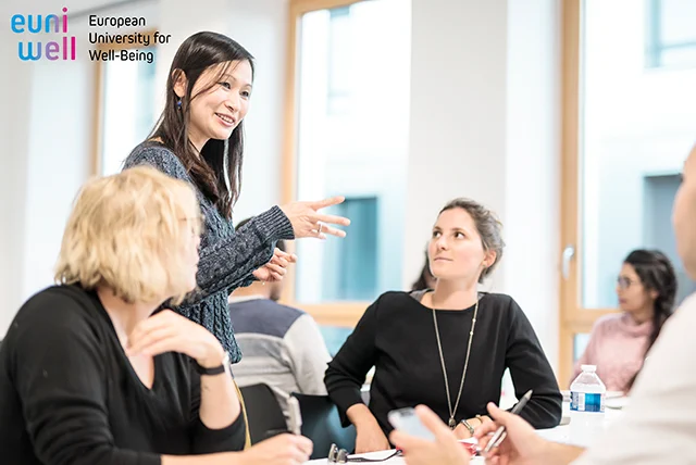 Women at a table in a study room engaged in a discussion. Logo: euniwell European University for Well-Being