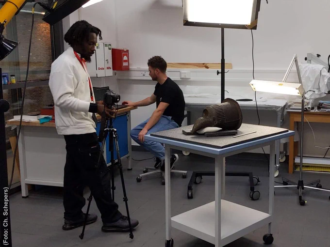 Foto, das zwei Mitarbeiter bei der Digitalisierung archäologischer Objekte zeigt. Auf einem kleinen Rolltisch liegt ein bronzener Gedenkkopf, links daneben steht ein Mann mit einer auf einem Stativ montierten Kamera. Im Hintergrund sitzt ein weiterer Mann an einem Computer. Mehrere Lampen leuchten das improvisierte Fotostudio aus.