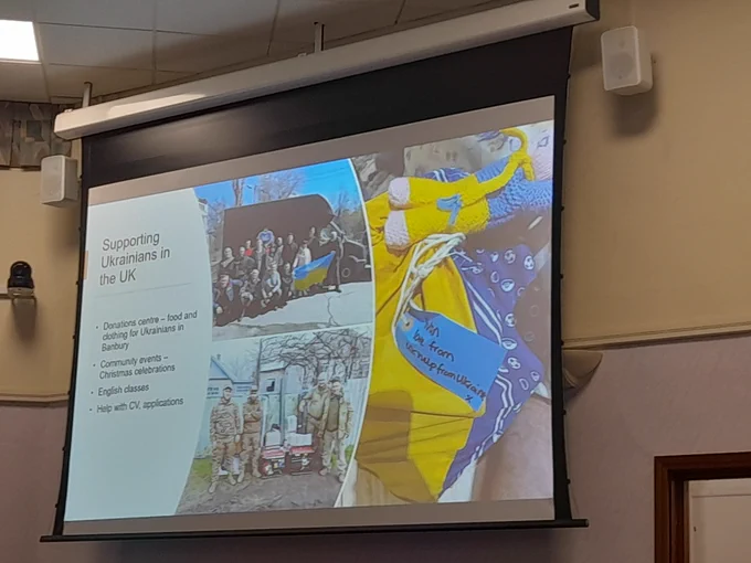 Slide projected onto a display screen. It reads 'Supporting Ukrainians in the UK. Donations centre - food and clothing for Ukrainians in Banabury. Community events - Christmas celebrations. English classes. Help with CV, applications.' With photos of aid deliveries to Ukraine.