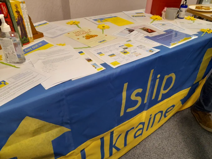 Information table for Islip Ukraine Community Support. The table is covered with a blue and yellow tablecloth with the words Islip and Ukraine. Information sheets, books and paper sunflowers cover the top of the table.