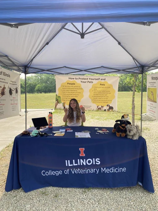Girl sitting behind a table labeled "Illinois College of Veterinary Medicine" under a tent, with signs hanging from the tent back and sides