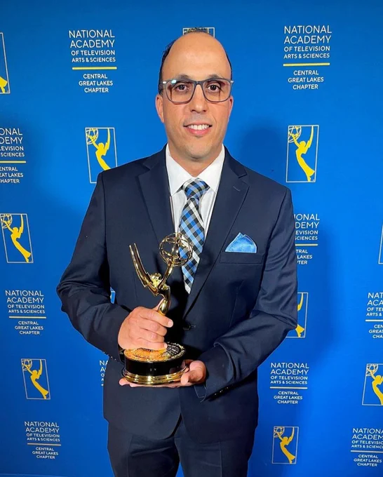 Saddam Al-Zubaidi poses in a navy suit, smiling with his emmy statue in hand. He stands in front of an all-blue backdrop with the Emmy logo. In gold lettering reads "National Academy of Television Arts & Sciences | Central Great Lakes Chapter"
