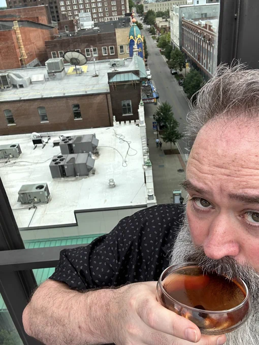 Bryce with a big beard and a Manhattan. He’s on a rooftop patio with the city in the background