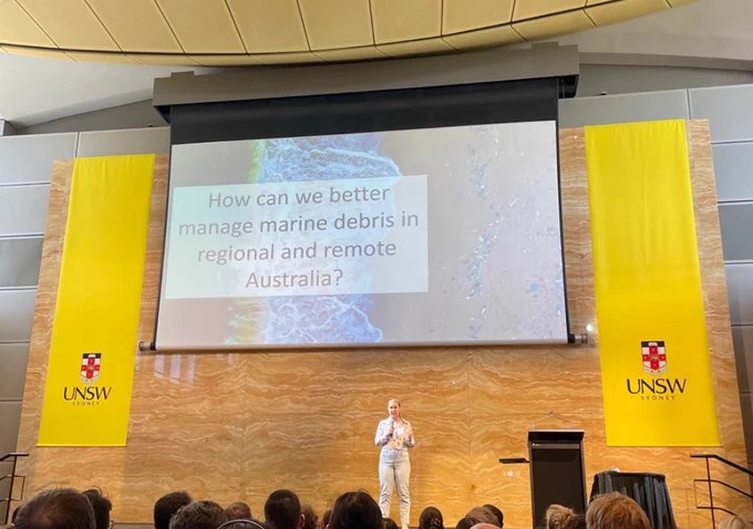 A scientist stands centrally on a stage, talking, mic in hand. Behind her, on screen, a slide reads "How can we better manage marine debris in regional and remote Australia?" 