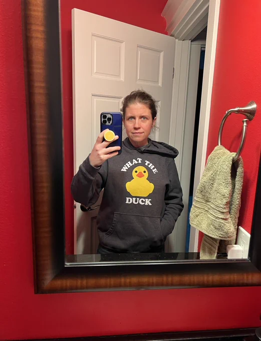 A woman taking a selfie in a mirror, wearing a hoodie with a yellow rubber ducky on it that says “WHAT THE DUCK”
