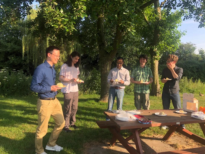 Photo of a group of people standing outside in the sunshine near a tree and a picnic table. Bo is holding a very big piece of cake and looks happy!