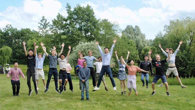 Photo of a large group of people standing in the sunshine in front of some trees jumping in the air and looking happy.