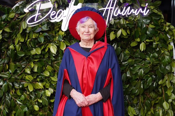 A photo of Deakin graduate Wendy, standing and smiling to camera in front of beautiful leaves and a sign saying ‘Deakin Alumni’