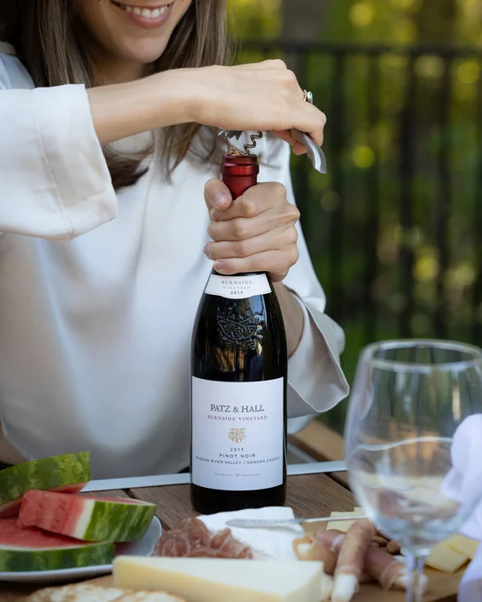 Women at an outdoor table opening a bottle of Patz & Hall Pinot Noir red wine. There is a cheese plate, wine glass and sliced watermelon on the table.