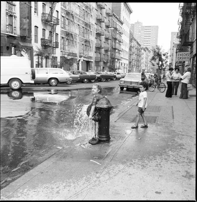 Another photograph of the two children playing in the hydrant. One of them catches water from the fire hydrant in her shirt.