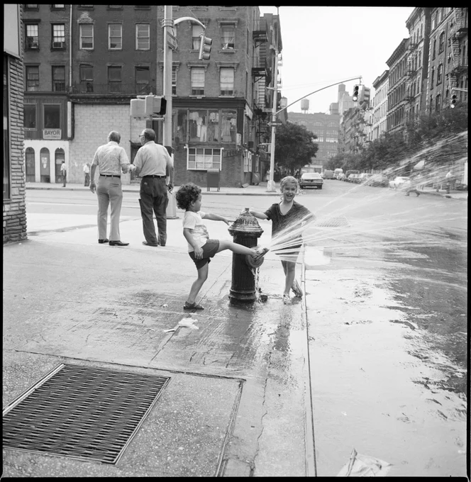 Two children play in an open fire hydrant. One is kicking a leg in the water. Another is smiling and looking right at the camera.