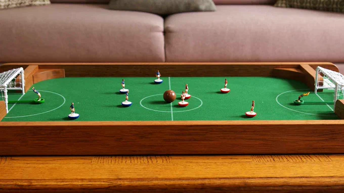 A FlickForKicks 5-a-side arena with meranti hardwood surrounds, sitting on a coffee table, with classic red and blue Subbuteo teams set up, ready for kicking-off the game.
