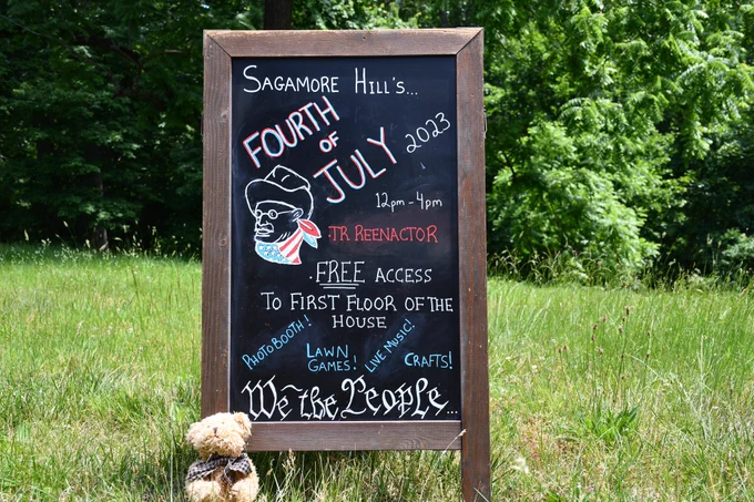 Chalkboard sign with teddy bear propped in front of it. Reads "Sagamore Hill's Fourth of July 2023, 12pm to 4pm. TR Reenactor, Free access to first floor of the house, photobooth, lawn games, live music, crafts, We the People written in the style of the constitution. 