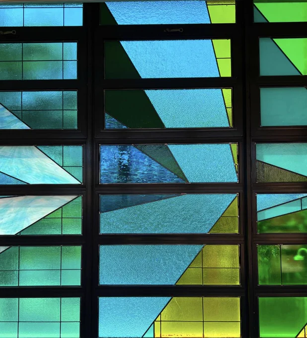 alt="A multi-colored stained glass window taken from the hallway of a school."  There are blues, yellow and green squares and triangles interspersed in an abstract pattern.