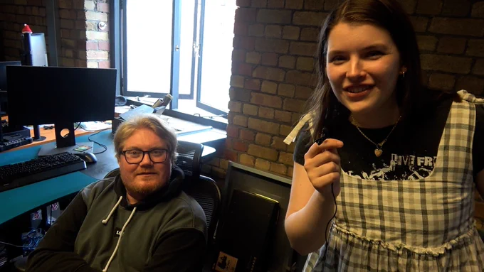 Sarah is looking at the camera holding a small microphone about to interview Duncan, who's looking at the camera, bewildered.