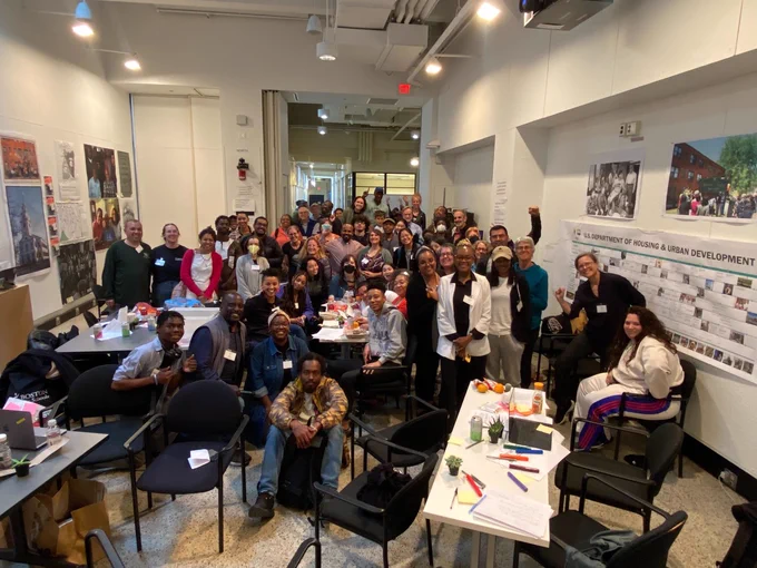 A diverse group of folks gathered in a room in building 7 at MIT, images of various posters/plots are hung on walls and desks and tables indicate where folks have been working together