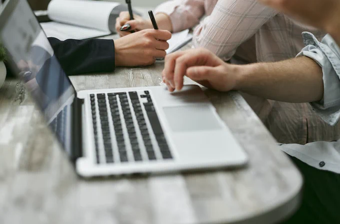 Crop faceless colleagues sitting at table while working on laptop and taking notes on documents with pen in light office