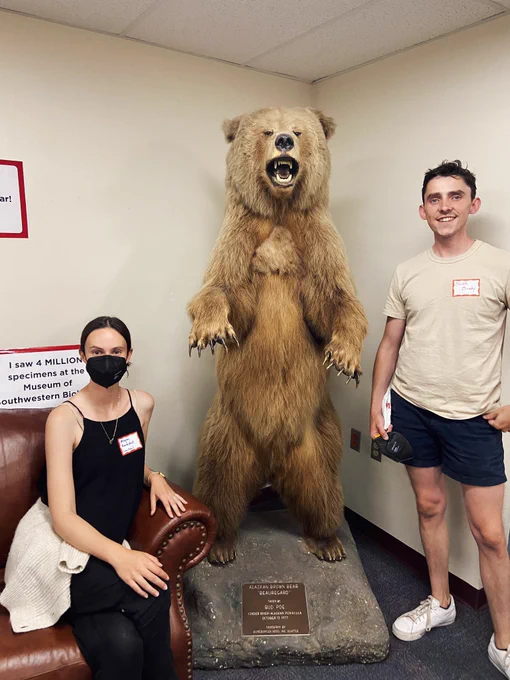 Megan, a brunette woman in a black mask, and Noah, a brunette man, are next to a standing, taxidermied brown bear.