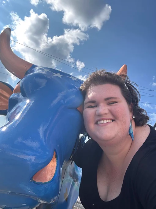 A woman takes a selfie and smiles while posing with one of the CowParade New England cows.