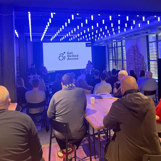 Live Nation employees are in a dark room seated on chairs and tables facing the large presentation screen. The screen is showing the Get Skilled Access logo. Either side of the screen, we have Get Skilled Access team presenting to the audience.