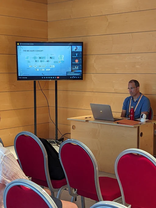 There is a man sitting in front of a. Computer in a conference room.  behind there is a screen showing the presentation of a speaker that is presenting online