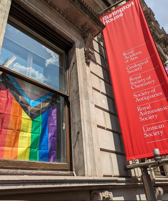 A progress Pride flag hanging proudly inside a wooden window frame in Burlington House, London, overlooking Piccadilly. A red banner to the right of the window lists the societies of Burlington House, include the Geological Society 