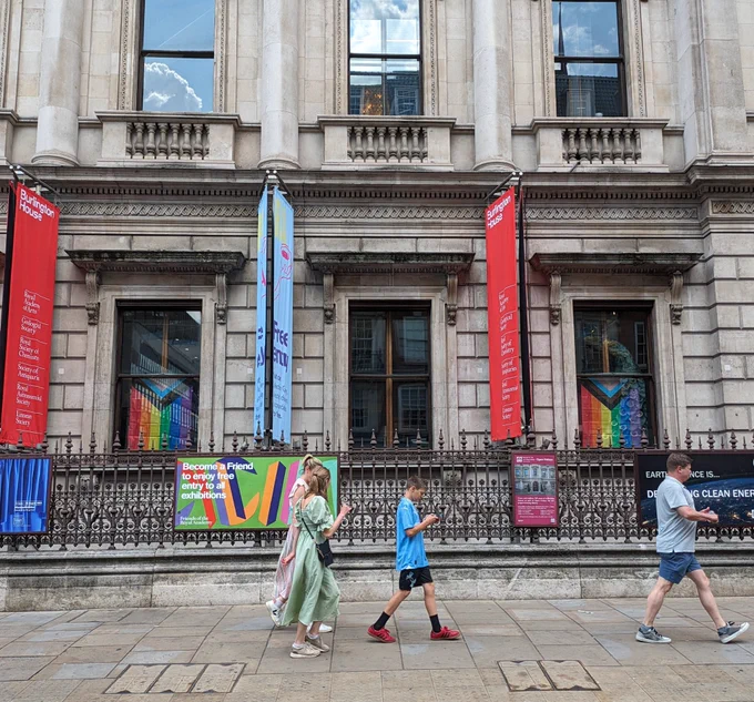 A photo of the front of the Geological Society at Burlington House looking into the Lower Library from the street (Piccadilly). Two progress Pride flags hang proudly in the windows 