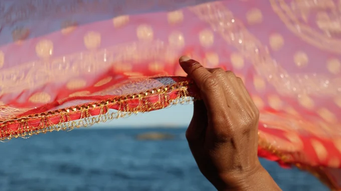  A photograph of a hand holding a sari that blows in the wind. The red and orange sari contrasts the blue water and sky of the horizon. 
