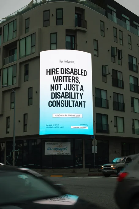 A three story billboard lit up on the corner of a building above a busy street. The billboard shows a white and blue gradient background with bold black text that reads: "Hey Hollywood, Hire Disabled Writers, Not Just A Disability Consultant."