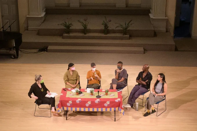 6 people sit in plastic chairs around a long rectangular table draped in a red table cloth with 3 small plants. One person speaks into a microphone.  