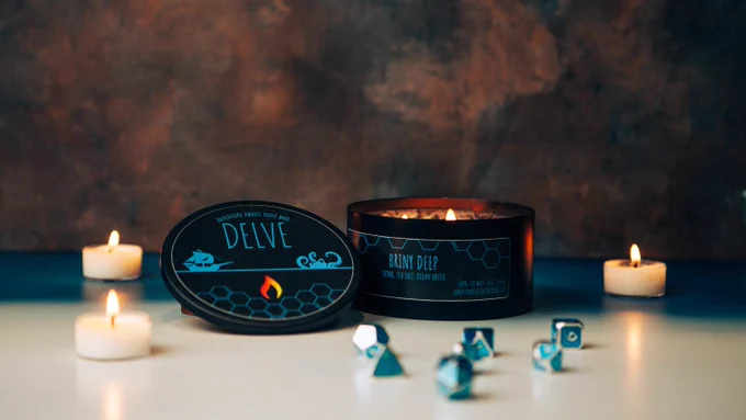 A lit candle with a black label reading "Briny Deep" in blue font. The central candle is surrounded by 3 lit tealights and the foreground has a metal set of polyhedral dice.