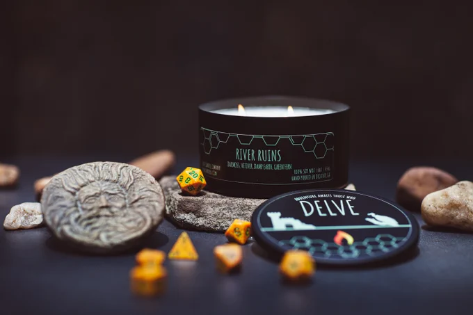 A lit candle with a black label reading "River Ruins" in light green font. The foreground has a yellow set of polyhedral dice.