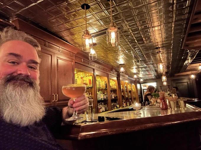 Bryce holding a cocktail with a big bushy beard in a speakeasy style bar sitting at the counter