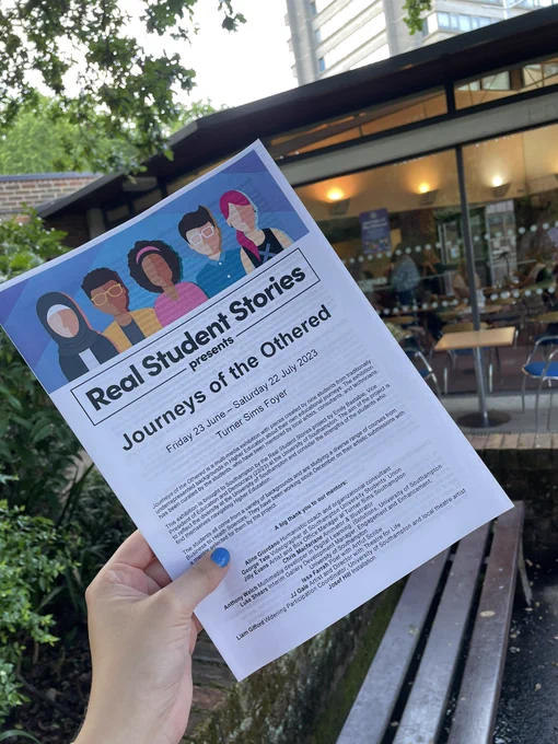 A hand holds a pamphlet for the exhibition. There is a lot of copy on the sheet, but we can clearly read ‘real student stories presents Journeys of the Othered’ at the top.