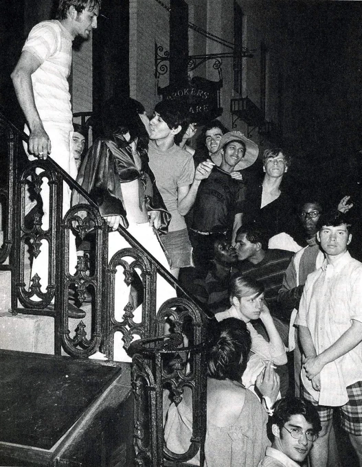 A group of young people gathered on a building’s steps near Stonewall, some looking into the camera and  smiling, two couples kissing