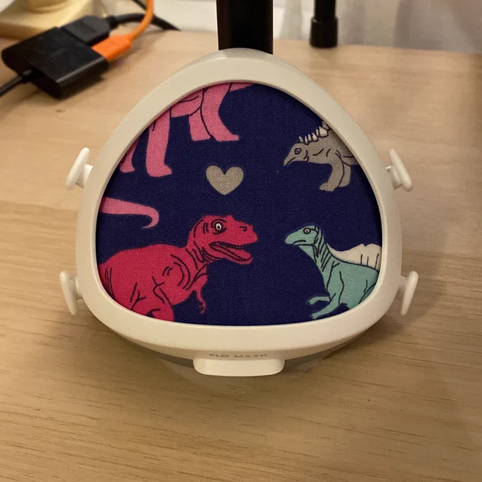 Flo mask without straps attached leaned against a post on a desk. The filter is covered with a fabric with brightly colored dinosaurs and hearts on a navy background.