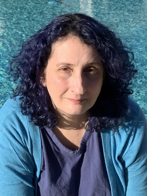 Poet JSA Lowe, a woman with indigo hair wearing a sky-blue sweater before a prismic cerulean pool.