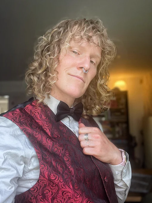 Huld, a white femme with a septum piercing and long blonde hair, poses wearing an ornate waistcoat and grey shirt with a black bow tie. This is the outfit of the character Jimmy Hall in the game At Dead of Night.