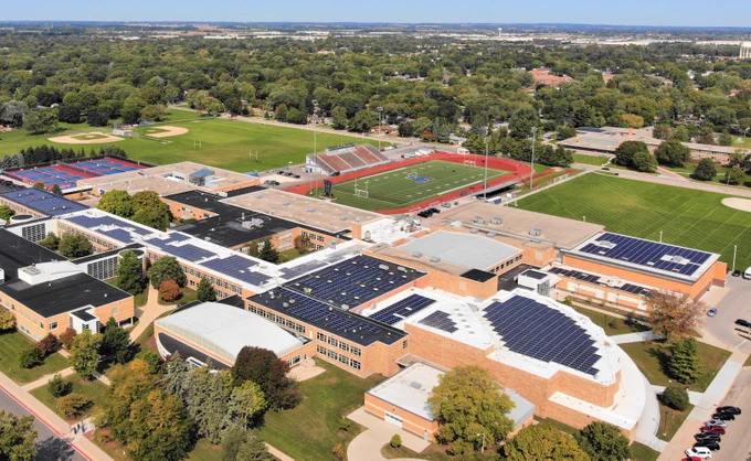 A drone photograph of West Aurora High School shows several solar arrays on multiple roof sections of the high school. The high school is nestled between a football field, tennis courts, baseball diamond, trees, and single-family homes. A few clouds appear but the sky is clear and blue. 
